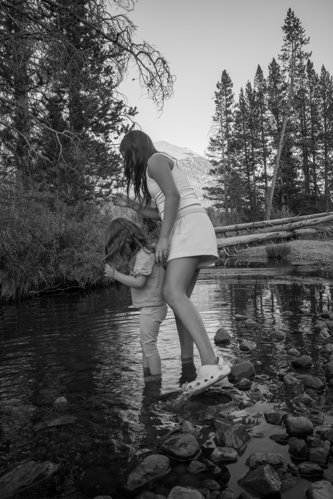 A woman steadies a child while they cross a shallow alpine stream in black and white.